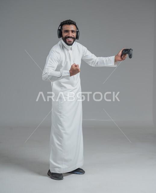 Modern entertaining technologies, having a fun and entertaining time, interacting and merging with PlayStation games, portrait of a Saudi Arabian Gulf man wearing traditional thobe, holding a joystick in his hand, wearing headphones and playing video games, gray background
