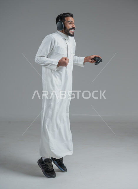 تقنيات حديثة مسلية, قضاء وقت ممتع ومسلي، التفاعل والاندماج مع ألعاب البلايستيشن، بورتريه لرجل عربي سعودي خليجي يرتدي الثوب التقليدي ويحمل بيده عصا التحكم  ويضع سماعات الرأس ويلعب بألعاب الفيديو، خلفية رمادية