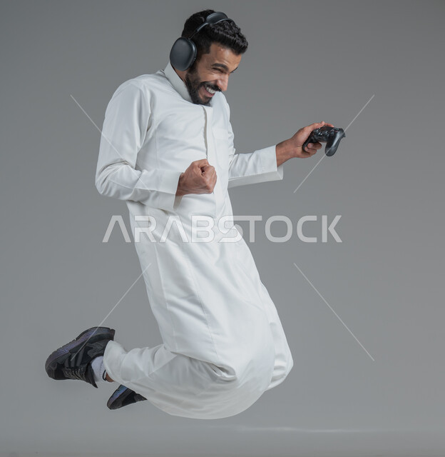 تقنيات حديثة مسلية, قضاء وقت ممتع ومسلي، التفاعل والاندماج مع ألعاب البلايستيشن، بورتريه لرجل عربي سعودي خليجي يرتدي الثوب التقليدي ويحمل بيده عصا التحكم  ويضع سماعات الرأس ويلعب بألعاب الفيديو، خلفية رمادية