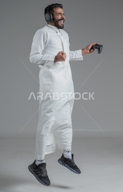 تقنيات حديثة مسلية, قضاء وقت ممتع ومسلي، التفاعل والاندماج مع ألعاب البلايستيشن، بورتريه لرجل عربي سعودي خليجي يرتدي الثوب التقليدي ويحمل بيده عصا التحكم  ويضع سماعات الرأس ويلعب بألعاب الفيديو، خلفية رمادية