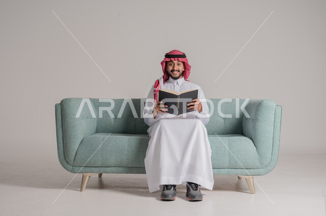 مفهوم محو الامية، قضاء وقت الفراغ بالتعلم والثقافة، حب المطالعة و الاستكشاف، بورتريه مقرب لرجل عربي سعودي خليجي يرتدي الشماغ والثوب التقليدي يجلس على الكنبة ويقرأ كتاب علمي تثقيفي، النظر الى الكاميرا بايماءات السعادة والسرور، خلفية رمادية