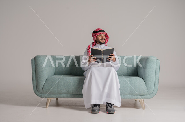 Love of reading and exploration, concept of literacy, close-up of a Saudi Arabian Gulf man wearing a traditional keffiyeh and thobe sitting on a sofa reading an educational scientific book, looking at something with gestures of happiness and pleasure, spending free time learning and culture, gray background