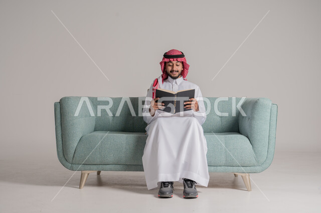 قضاء وقت الفراغ بالتعلم والثقافة، حب المطالعة و الاستكشاف، مفهوم محو الامية، بورتريه مقرب لرجل عربي سعودي خليجي يرتدي الشماغ والثوب التقليدي يجلس على الكنبة ويقرأ كتاب علمي تثقيفي، القراءة بإيماءات التمعن والتركيز، خلفية رمادية