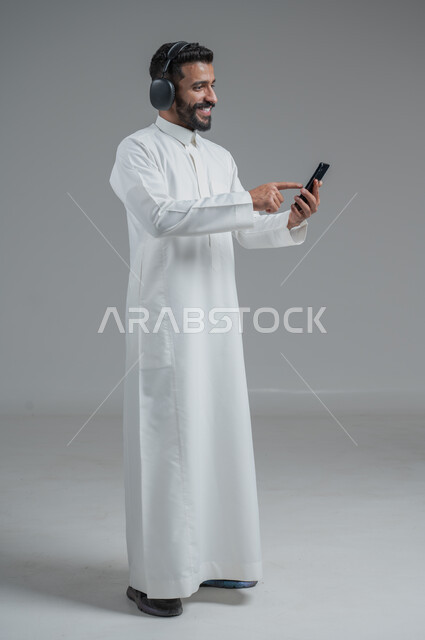 الدردشة والتواصل مع الأصدقاء عبر الجوال, استخدام التكنولوجيا الحديثة و المتطورة, بورتريه لرجل عربي خليجي سعودي يرتدي الثوب التقليدي  يمسك الهاتف المحمول بيده و يضع سماعات الرأس, صورة الجسم بالطول الكامل, خلفية رمادية