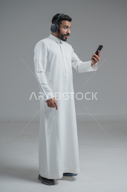 الدردشة والتواصل مع الأصدقاء عبر الجوال, استخدام التكنولوجيا الحديثة و المتطورة, بورتريه لرجل عربي خليجي سعودي يرتدي الثوب التقليدي  يمسك الهاتف المحمول بيده و يضع سماعات الرأس, صورة الجسم بالطول الكامل, خلفية رمادية