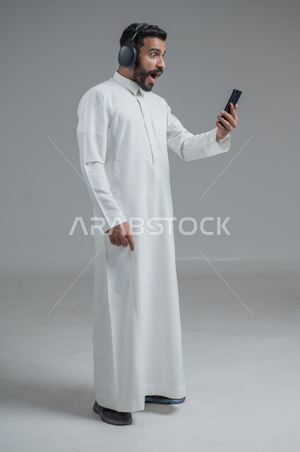 الدردشة والتواصل مع الأصدقاء عبر الجوال, استخدام التكنولوجيا الحديثة و المتطورة, بورتريه لرجل عربي خليجي سعودي يرتدي الثوب التقليدي  يمسك الهاتف المحمول بيده و يضع سماعات الرأس, صورة الجسم بالطول الكامل, خلفية رمادية