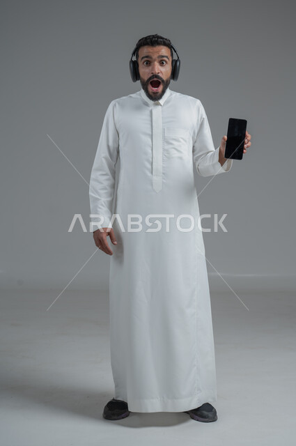 الدردشة والتواصل مع الأصدقاء عبر الجوال, استخدام التكنولوجيا الحديثة و المتطورة, بورتريه لرجل عربي خليجي سعودي يرتدي الثوب التقليدي  يمسك الهاتف المحمول بيده و يضع سماعات الرأس, صورة الجسم بالطول الكامل, خلفية رمادية