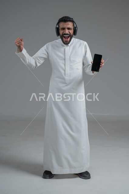 الدردشة والتواصل مع الأصدقاء عبر الجوال, استخدام التكنولوجيا الحديثة و المتطورة, بورتريه لرجل عربي خليجي سعودي يرتدي الثوب التقليدي  يمسك الهاتف المحمول بيده و يضع سماعات الرأس, صورة الجسم بالطول الكامل, خلفية رمادية