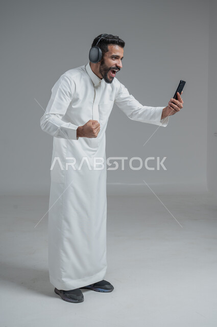 الدردشة والتواصل مع الأصدقاء عبر الجوال, استخدام التكنولوجيا الحديثة و المتطورة, بورتريه لرجل عربي خليجي سعودي يرتدي الثوب التقليدي  يمسك الهاتف المحمول بيده و يضع سماعات الرأس, صورة الجسم بالطول الكامل, خلفية رمادية