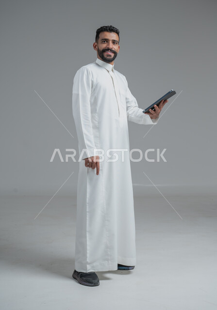 استخدام التكنولوجيا والأجهزة التقنية الحديثة المتطورة، ايماءات السعادة والفرح، بورتريه  لرجل عربي خليجي سعودي مبتسم يرتدي الثوب التقليدي يمسك بيده الهاتف المحمول، خلفية رمادية