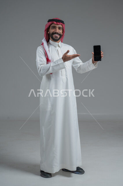 استخدام التكنولوجيا والأجهزة التقنية الحديثة المتطورة، ايماءات السعادة والفرح، بورتريه  لرجل عربي خليجي سعودي مبتسم يرتدي الثوب التقليدي يمسك بيده الهاتف المحمول، خلفية رمادية