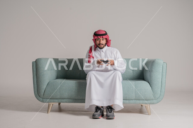 تعابير الاستمتاع باللعب بالألعاب الالكترونية الترفيهية، استخدام الأجهزة التكنولوجية والتقنيات الحديثة، بورتريه لرجل عربي خليجي سعودي مبتسم يرتدي الثوب التقليدي والشماغ يجلس على الكنبة ويمسك بيده عصا التحكم بإيماءات السرور والاندماج، خلفية رمادية