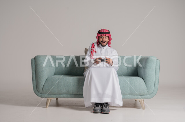 استخدام الأجهزة التكنولوجية والتقنيات الحديثة، بورتريه لرجل عربي خليجي سعودي مبتسم يرتدي الثوب التقليدي والشماغ يجلس على الكنبة ويمسك بيده عصا التحكم بإيماءات السرور والاندماج، تعابير الاستمتاع باللعب بالألعاب الالكترونية الترفيهية، خلفية رمادية