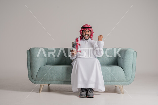 المتعة والمرح بألعاب الفيديو والبلايستيشن الترفيهية , بورتريه لرجل عربي خليجي سعودي يرتدي الثوب التقليدي والشماغ يجلس على الكنبة ويمسك بيده عصا التحكم، استخدام الأجهزة التكنولوجية والتقنيات الحديثة، إيماءات وجه ويدين تدل على الفوز و الانتصار، خلفية رمادية