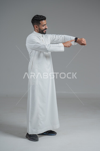 تيسير وتسهيل المهام اليومية باستخدام أحدث التقنيات, تتبع الإشعارات عبر الساعات الحديثة, بورتريه  لرجل عربي خليجي سعودي يرتدي الثوب التقليدي ويلبس في يده ساعة ذكية، الجسم بالطول الكامل، خلفية رمادية