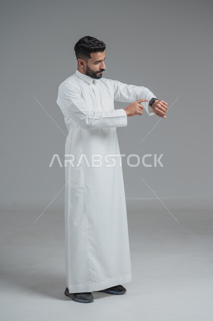 تيسير وتسهيل المهام اليومية باستخدام أحدث التقنيات, تتبع الإشعارات عبر الساعات الحديثة, بورتريه  لرجل عربي خليجي سعودي يرتدي الثوب التقليدي ويلبس في يده ساعة ذكية، الجسم بالطول الكامل، خلفية رمادية