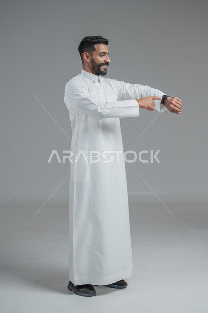 تيسير وتسهيل المهام اليومية باستخدام أحدث التقنيات, تتبع الإشعارات عبر الساعات الحديثة, بورتريه  لرجل عربي خليجي سعودي يرتدي الثوب التقليدي ويلبس في يده ساعة ذكية، الجسم بالطول الكامل، خلفية رمادية