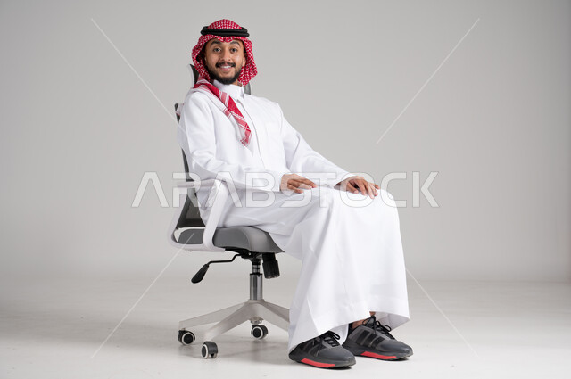 النظر إلى الكاميرا بإيماءات السعادة والسرور، الاهتمام بالمظهر الخارجي والثقة بالنفس، بورتريه لرجل عربي خليجي سعودي يرتدي الشماغ والثوب التقليدي يجلس على كرسي دوار بهدوء واتزان، مفهوم الأناقة والرجولة، الجسم بالطول الكامل، خلفية رمادية