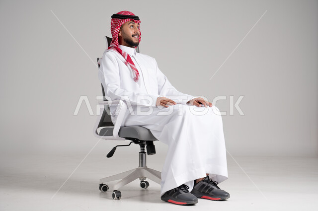 الاهتمام بالمظهر الخارجي والثقة بالنفس، بورتريه لرجل عربي خليجي سعودي يرتدي الشماغ والثوب التقليدي يجلس على كرسي دوار بهدوء واتزان وينظر إلى مكان ما بإيماءات التفكير العميق، مفهوم الأناقة والرجولة، الجسم بالطول الكامل، خلفية رمادية