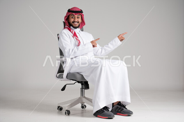 الاهتمام بالمظهر الخارجي والثقة بالنفس، حركات وجه ويدين تشير إلى المفاجأة، بورتريه لرجل عربي خليجي سعودي يرتدي الشماغ والثوب التقليدي يجلس على كرسي دوار ويرفع يديه باتجاه محدد بإيماءات الدهشة، مفهوم الأناقة والرجولة، الجسم بالطول الكامل، خلفية رمادية