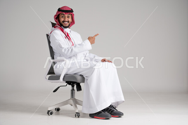 الإشارة باليد نحو مكان ما بعيد، بورتريه لرجل عربي خليجي سعودي يرتدي الشماغ والثوب التقليدي يجلس على كرسي دوار ويرفع إصبعه باتجاه محدد بإيماءات السرور، مفهوم الأناقة والرجولة، الاهتمام بالمظهر الخارجي والثقة بالنفس، الجسم بالطول الكامل، خلفية رمادية