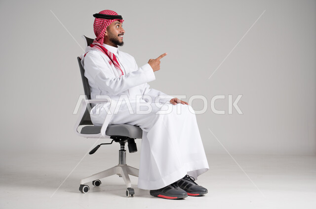 الإشارة باليد نحو مكان ما بعيد، بورتريه لرجل عربي خليجي سعودي يرتدي الشماغ والثوب التقليدي يجلس على كرسي دوار ويرفع إصبعه باتجاه محدد بإيماءات السرور، مفهوم الأناقة والرجولة، الاهتمام بالمظهر الخارجي والثقة بالنفس، الجسم بالطول الكامل، خلفية رمادية