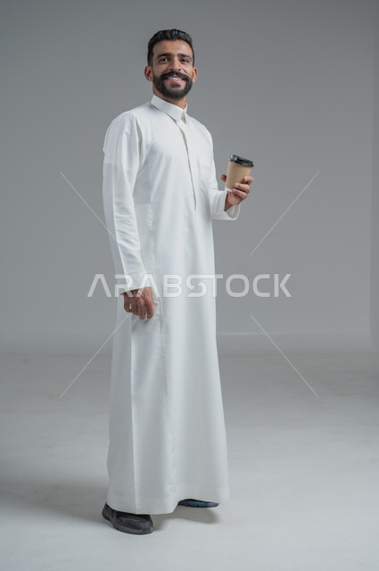 Effect of caffeine on brain activity and memory, enjoying hot drinks and warm winter herbs, portrait of a Saudi Arabian Gulf man wearing traditional thobe holding a cardboard cup of coffee, full body image, gray background
