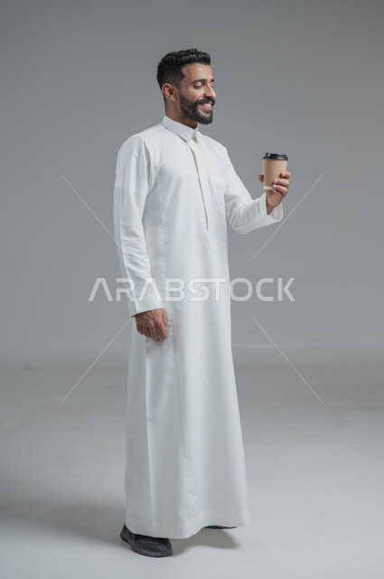 Effect of caffeine on brain activity and memory, enjoying hot drinks and warm winter herbs, portrait of a Saudi Arabian Gulf man wearing traditional thobe holding a cardboard cup of coffee, full body image, gray background