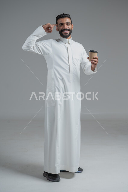 تأثير الكافيين على نشاط الدماغ والذاكرة، الاستمتاع بتناول المشروبات الساخنة والأعشاب الشتوية الدافئة، بورتريه لرجل عربي خليجي سعودي يرتدي الثوب التقليدي يحمل بيده كوب ورقي مقوى من القهوة، صورة الجسم بالطول الكامل، خلفية رمادية