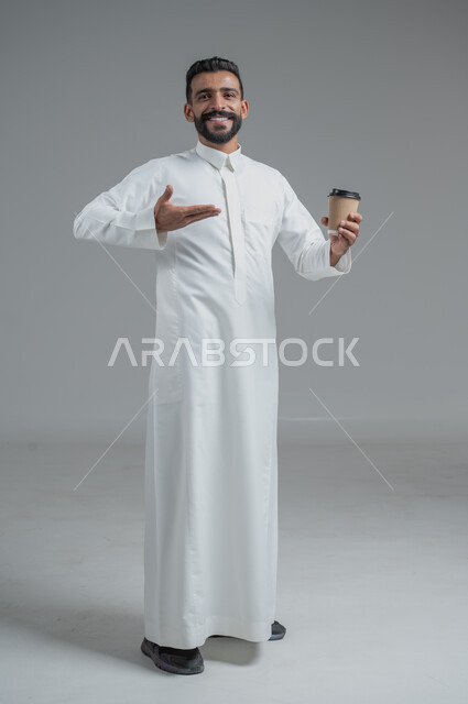 تأثير الكافيين على نشاط الدماغ والذاكرة، الاستمتاع بتناول المشروبات الساخنة والأعشاب الشتوية الدافئة، بورتريه لرجل عربي خليجي سعودي يرتدي الثوب التقليدي يحمل بيده كوب ورقي مقوى من القهوة، صورة الجسم بالطول الكامل، خلفية رمادية