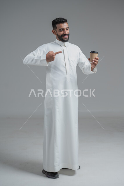 تأثير الكافيين على نشاط الدماغ والذاكرة، الاستمتاع بتناول المشروبات الساخنة والأعشاب الشتوية الدافئة، بورتريه لرجل عربي خليجي سعودي يرتدي الثوب التقليدي يحمل بيده كوب ورقي مقوى من القهوة، صورة الجسم بالطول الكامل، خلفية رمادية