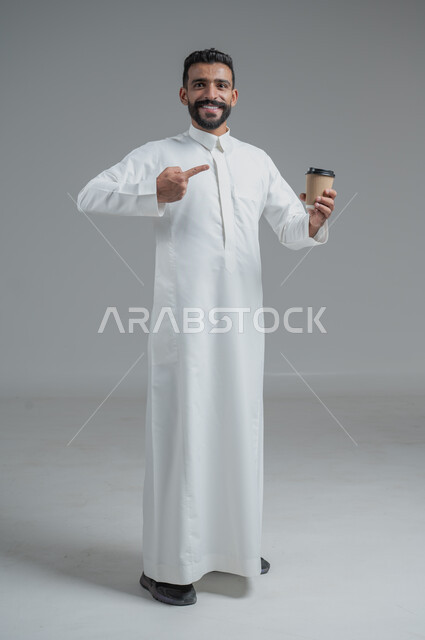 تأثير الكافيين على نشاط الدماغ والذاكرة، الاستمتاع بتناول المشروبات الساخنة والأعشاب الشتوية الدافئة، بورتريه لرجل عربي خليجي سعودي يرتدي الثوب التقليدي يحمل بيده كوب ورقي مقوى من القهوة، صورة الجسم بالطول الكامل، خلفية رمادية