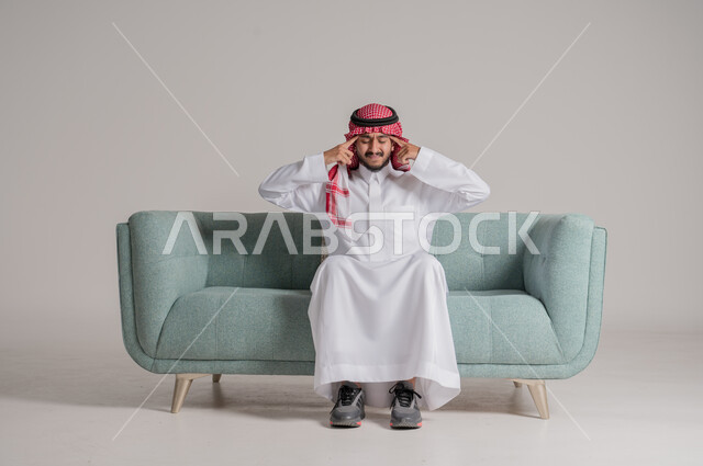 الحاجة إلى العلاج والمساعده, بورتريه لرجل عربي خليجي سعودي يرتدي الثوب التقليدي والشماغ يجلس على الكنبه ويضع يديه على رأسه بإيماءات الانزعاج والتعب, الشعور بالمرض وألم الرأس, آلام الشقيقة والصداع النصفي, خلفية رمادية