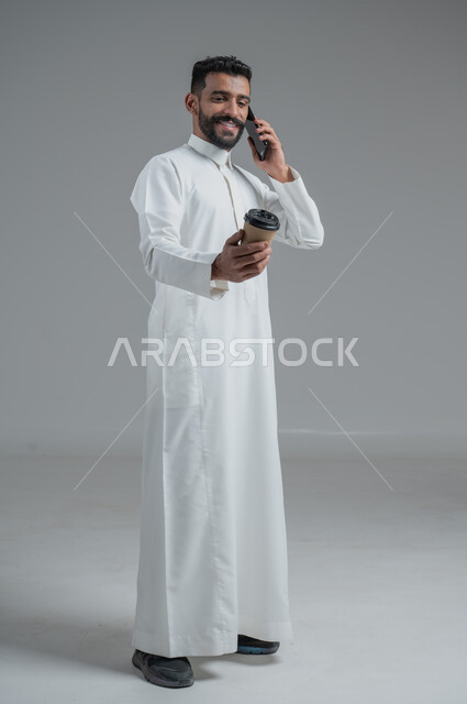 الاستمتاع بتناول المشروبات الساخنة والأعشاب الشتوية الدافئة، تأثير الكافيين على نشاط الدماغ والذاكرة، بورتريه لرجل عربي خليجي سعودي يرتدي الثوب التقليدي يحمل بيده كوب ورقي مقوى من القهوة ويستخدم الجهاز المحمول، صورة الجسم بالطول الكامل، خلفية رمادية