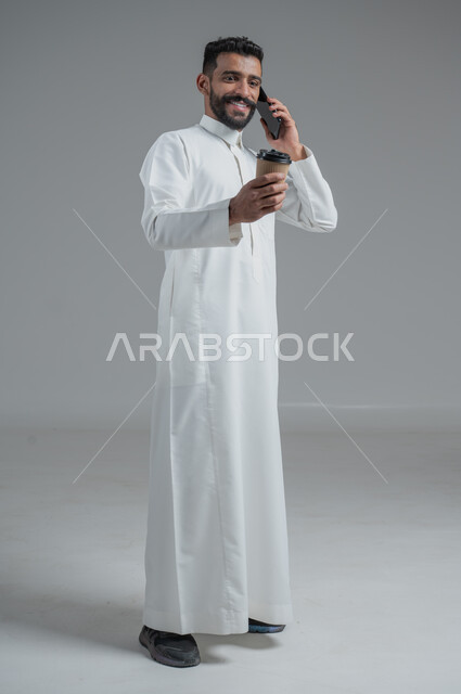 الاستمتاع بتناول المشروبات الساخنة والأعشاب الشتوية الدافئة، تأثير الكافيين على نشاط الدماغ والذاكرة، بورتريه لرجل عربي خليجي سعودي يرتدي الثوب التقليدي يحمل بيده كوب ورقي مقوى من القهوة ويستخدم الجهاز المحمول، صورة الجسم بالطول الكامل، خلفية رمادية