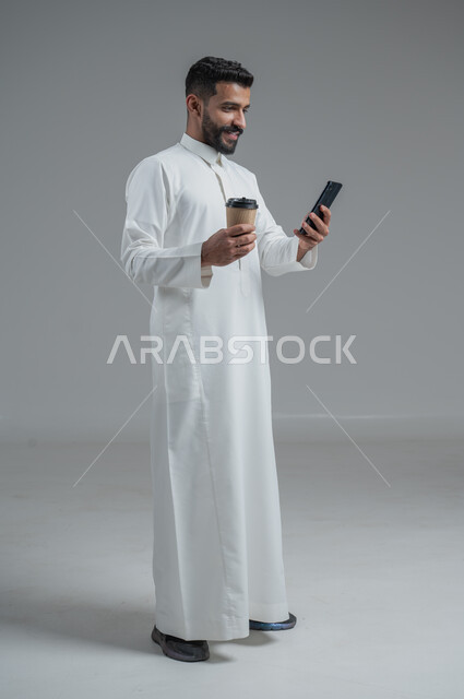 الاستمتاع بتناول المشروبات الساخنة والأعشاب الشتوية الدافئة، تأثير الكافيين على نشاط الدماغ والذاكرة، بورتريه لرجل عربي خليجي سعودي يرتدي الثوب التقليدي يحمل بيده كوب ورقي مقوى من القهوة ويستخدم الجهاز المحمول، صورة الجسم بالطول الكامل، خلفية رمادية