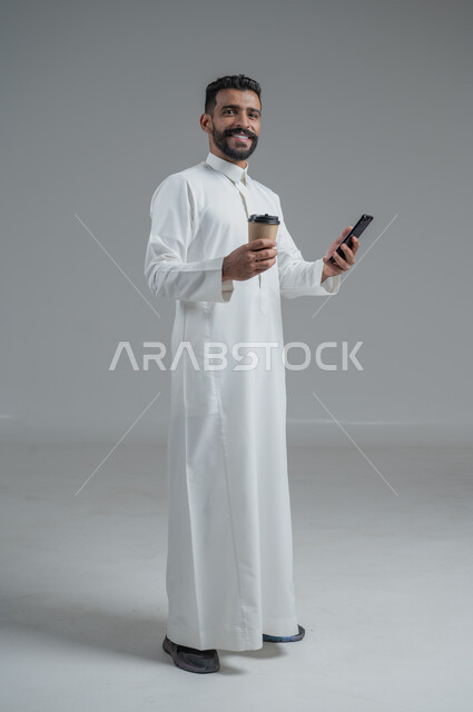 الاستمتاع بتناول المشروبات الساخنة والأعشاب الشتوية الدافئة، تأثير الكافيين على نشاط الدماغ والذاكرة، بورتريه لرجل عربي خليجي سعودي يرتدي الثوب التقليدي يحمل بيده كوب ورقي مقوى من القهوة ويستخدم الجهاز المحمول، صورة الجسم بالطول الكامل، خلفية رمادية