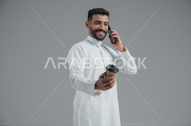 الاستمتاع بتناول المشروبات الساخنة والأعشاب الشتوية الدافئة، تأثير الكافيين على نشاط الدماغ والذاكرة، بورتريه مقرب لرجل عربي خليجي سعودي يرتدي الثوب التقليدي يحمل بيده كوب ورقي مقوى من القهوة ويستخدم الجهاز المحمول، خلفية رمادية