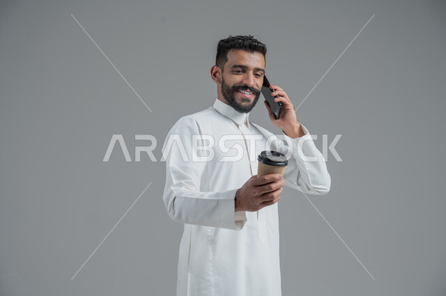 الاستمتاع بتناول المشروبات الساخنة والأعشاب الشتوية الدافئة، تأثير الكافيين على نشاط الدماغ والذاكرة، بورتريه مقرب لرجل عربي خليجي سعودي يرتدي الثوب التقليدي يحمل بيده كوب ورقي مقوى من القهوة ويستخدم الجهاز المحمول، خلفية رمادية