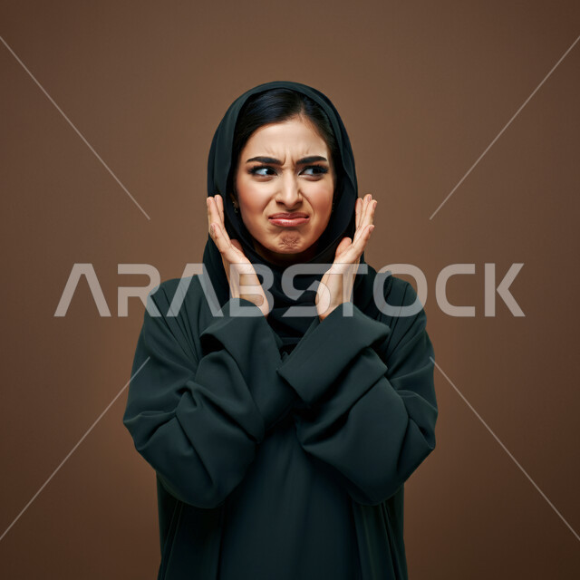 تعابير الغضب والعصبية، بورتريه مقرب لإمرأة عربيه خليجية سعودية محجبة ترتدي العباءة السوداء ترفع يديها للأعلى لعدم معرفة أمر ما, النظر الى مكان ما بإيماءات العبوس والاشمئزاز، الشعور بالإنزعاج وعدم الرغبة والقبول, خلفية بنية
