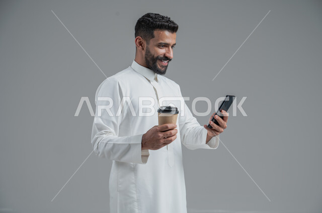 الاستمتاع بتناول المشروبات الساخنة والأعشاب الشتوية الدافئة، تأثير الكافيين على نشاط الدماغ والذاكرة، بورتريه مقرب لرجل عربي خليجي سعودي يرتدي الثوب التقليدي يحمل بيده كوب ورقي مقوى من القهوة ويستخدم الجهاز المحمول، خلفية رمادية