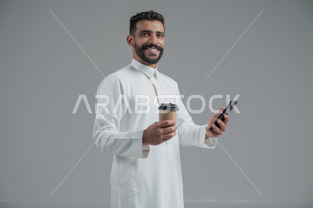 الاستمتاع بتناول المشروبات الساخنة والأعشاب الشتوية الدافئة، تأثير الكافيين على نشاط الدماغ والذاكرة، بورتريه مقرب لرجل عربي خليجي سعودي يرتدي الثوب التقليدي يحمل بيده كوب ورقي مقوى من القهوة ويستخدم الجهاز المحمول، خلفية رمادية