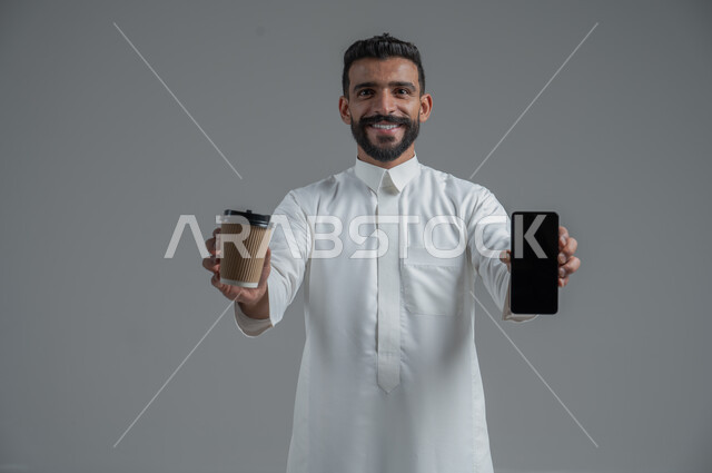 الاستمتاع بتناول المشروبات الساخنة والأعشاب الشتوية الدافئة، تأثير الكافيين على نشاط الدماغ والذاكرة، بورتريه مقرب لرجل عربي خليجي سعودي يرتدي الثوب التقليدي يحمل بيده كوب ورقي مقوى من القهوة ويستخدم الجهاز المحمول، خلفية رمادية