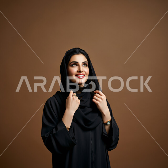 تعابير البهجة والسرور والسعادة، الاهتمام بالأناقة والمظهر الخارجي، مفهوم الأنوثة والنعومة، بورتريه مقرب لامرأة عربية خليجية سعودية محجبة مبتسمة ترتدي العباءة السوداء تنظر إلى الأعلى  بإيماءات الثقة بالنفس والفرح، خلفية بنية