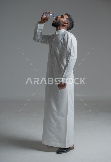 أهمية شرب الماء النقي للمحافظة على بشرة نضرة، بورتريه لرجل عربي خليجي سعودي يرتدي الثوب التقليدي ويشرب المياه من عبوة بلاستيكية، المحافظة على سلامة الجهاز المناعي، التخلص من سموم الجسم، الحفاظ على صحة الكلى ومعالجتها، الجسم بالطول الكامل، خلفية رمادية