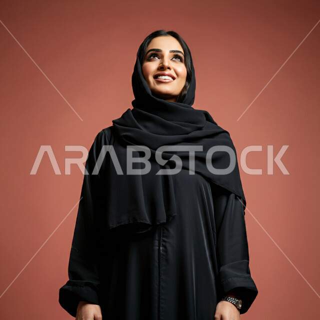 الاهتمام بالأناقة والمظهر الخارجي، مفهوم الأنوثة والنعومة، بورتريه مقرب لامرأة عربية خليجية سعودية محجبة مبتسمة ترتدي العباءة السوداء تنظر إلى الأعلى  بإيماءات الثقة بالنفس والفرح، تعابير السرور والسعادة، خلفية حمراء