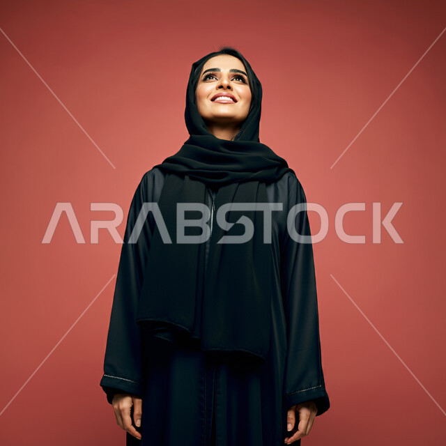  الاهتمام بالأناقة والمظهر الخارجي، مفهوم الأنوثة والنعومة، بورتريه مقرب لامرأة عربية خليجية سعودية محجبة مبتسمة ترتدي العباءة السوداء تنظر إلى الأعلى  بإيماءات الثقة بالنفس والفرح، تعابير السرور والسعادة، خلفية حمراء