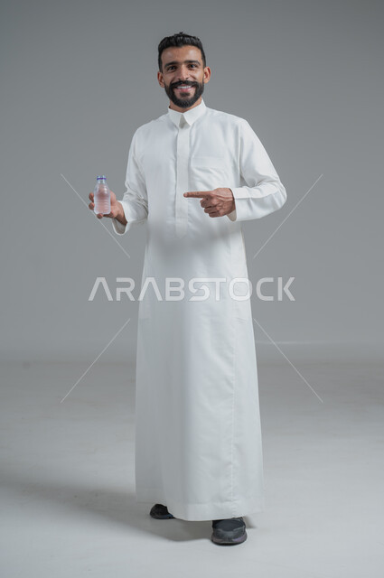 المحافظة على سلامة الجهاز المناعي، التخلص من سموم الجسم، الحفاظ على صحة الكلى ومعالجتها، أهمية شرب الماء النقي للمحافظة على بشرة نضرة، بورتريه لرجل عربي خليجي سعودي يرتدي الثوب التقليدي ويمسك عبوة بلاستيكية من المياه، الجسم بالطول الكامل، خلفية رمادية
