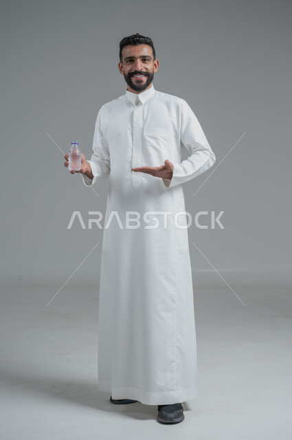 المحافظة على سلامة الجهاز المناعي، التخلص من سموم الجسم، الحفاظ على صحة الكلى ومعالجتها، أهمية شرب الماء النقي للمحافظة على بشرة نضرة، بورتريه لرجل عربي خليجي سعودي يرتدي الثوب التقليدي ويمسك عبوة بلاستيكية من المياه، الجسم بالطول الكامل، خلفية رمادية