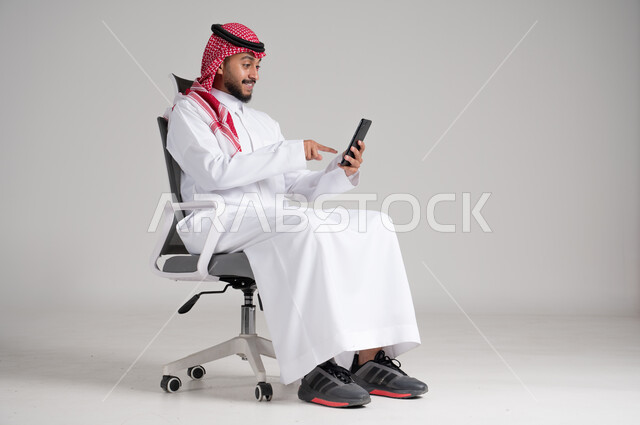 استخدام الأجهزة والتقنيات الحديثة، بورتريه لرجل عربي خليجي سعودي يرتدي الشماغ والثوب التقليدي يجلس على كرسي دوار يمسك الجوال بيده ويشير إليه بإيماءات السرور، مفهوم الأناقة والرجولة، الاهتمام بالمظهر الخارجي والثقة بالنفس، الجسم بالطول الكامل، خلفية رمادية